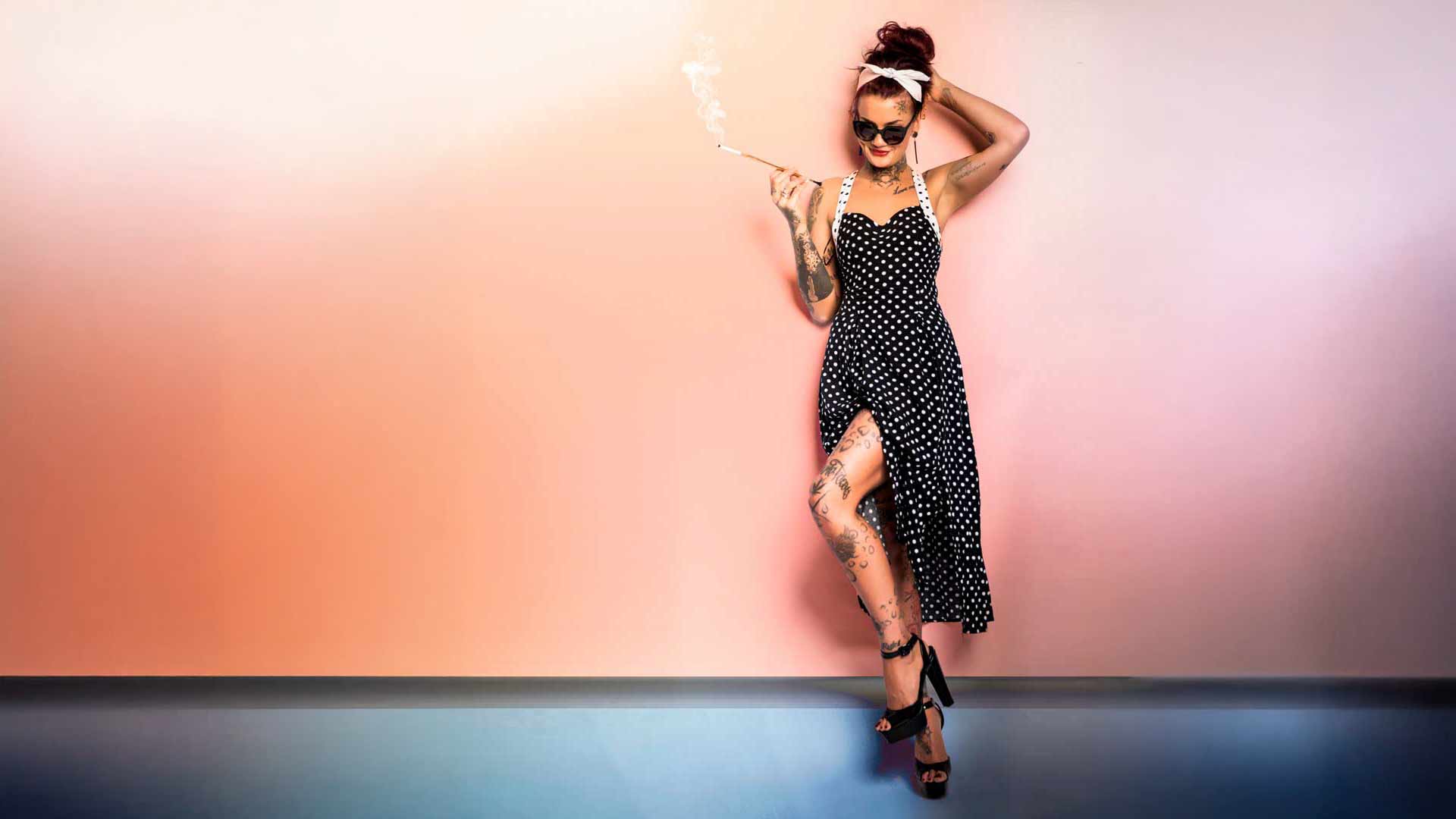 Ganzkörperaufnahme einer Frau im Vintage-Pin-up-Look: schwarzes Neckholder-Kleid mit weißen Punkten und Beinschlitz, Tattoos an Armen und Beinen, Sonnenbrille, Kopftuch und Zigarettenspitze, vor einer pastellrosa Wand mit dunklerem Bodenbereich