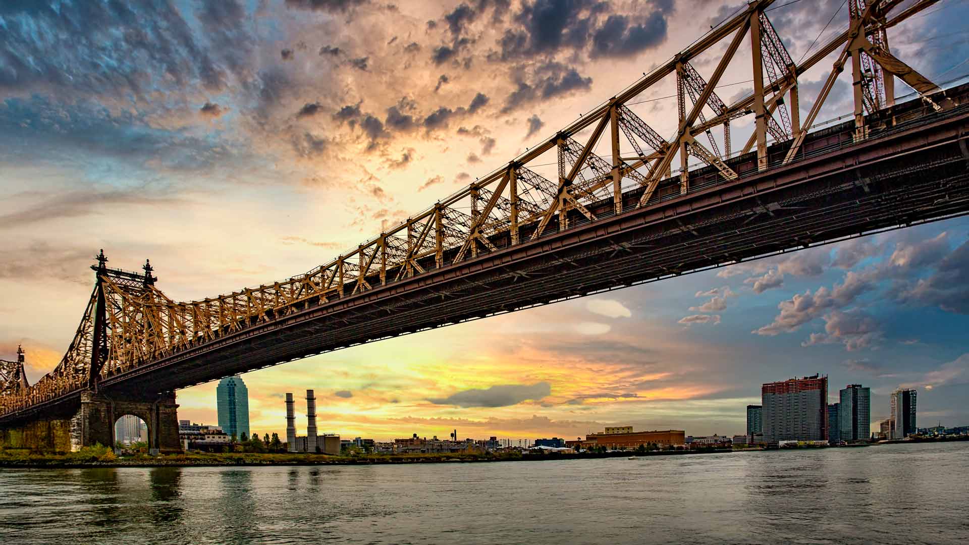 Queensboro Bridge bei Sonnenuntergang: Eine atemberaubende Ansicht der iconic Queensboro Bridge, die sich über den East River erstreckt, mit der Skyline von Manhattan und Long Island City im Hintergrund, alles getaucht in die warmen Farbtöne eines Sonnenuntergangs.