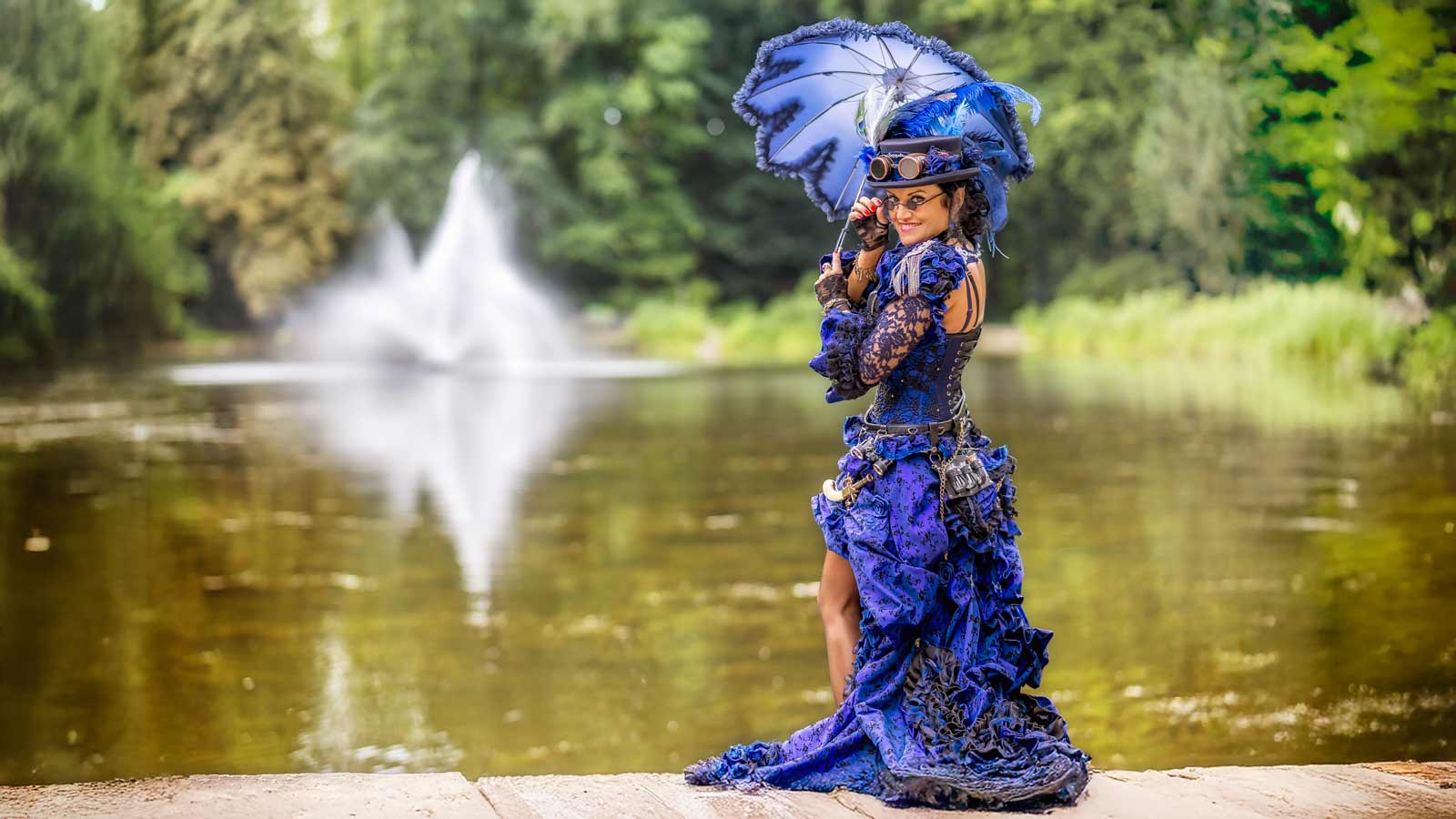 Frau in blauem Steampunk-Kleid mit Schirm am See.