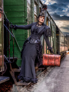 Eine Frau in einem schwarzen, gestreiften, viktorianisch/Steampunk-inspirierten Kleid mit Korsett und Hut, die mit einem braunen Vintage-Koffer die Stufen eines grünen Eisenbahnwaggons betritt.