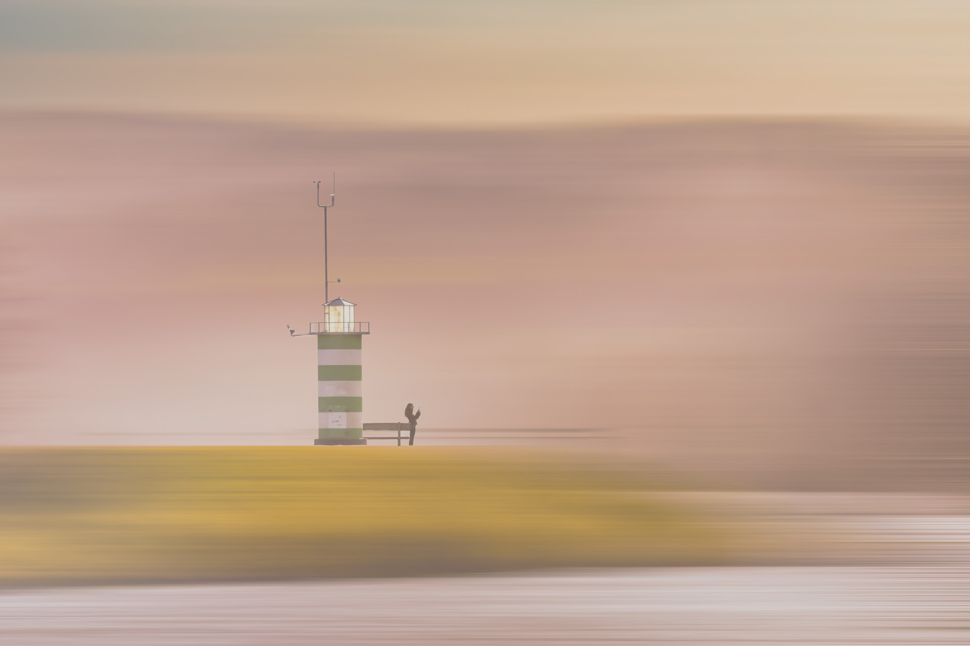 „Grün-weiß gestreifter Leuchtturm mit Person am Fuß vor pastellfarbenem Himmel bei Sonnenuntergang