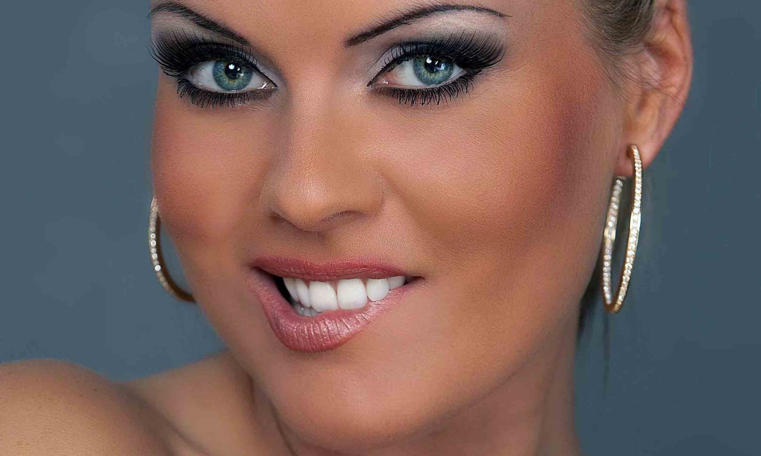 Frau mit starkem Augen Make-up, langen Wimpern und großen funkelnden Creolen lächelt in die Kamera – Beauty Close-up Portrait vor grauem Hintergrund.