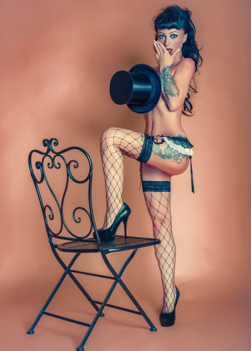 Dieses Bild zeigt eine Pin-up-Frau im Stil der Burlesque, die eine provokative Pose einnimmt. Sie trägt schwarze Netzstrümpfe mit Spitzenbändern und schwarze High Heels. Sie hält einen schwarzen Zylinderhut kokett vor ihrem Körper und ihre andere Hand bedeckt leicht ihren Mund. Sie hat sichtbare Tätowierungen auf dem Arm und Oberschenkel. Ihr linkes Bein ist auf einem schwarzen, verschnörkelten Metallstuhl platziert, der sich auf der linken Seite des Bildes befindet. Der Hintergrund ist ein einfarbiger, heller Braunton, der die Aufmerksamkeit auf die Frau lenkt. Die Beleuchtung betont ihre Figur und die Details ihrer Kleidung.