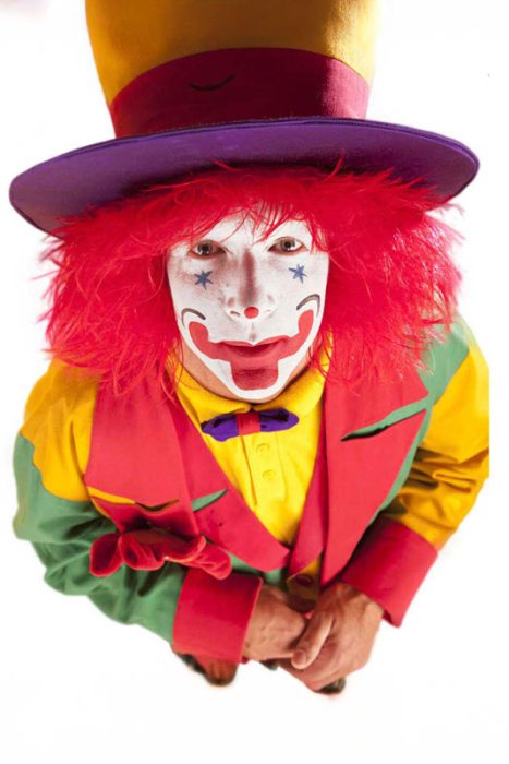 Nahaufnahme eines lächelnden Clowns mit roter Perücke, buntem Kostüm und Make-up, der einen Zylinder trägt und in die Kamera schaut.