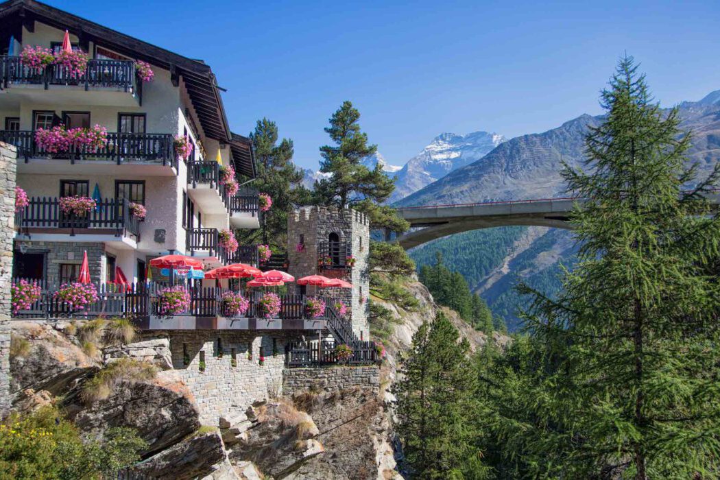Blumengeschmücktes Alpenhotel mit Burgmauer und Terrasse mit roten Sonnenschirmen hoch über einer Schlucht, im Hintergrund Brücke und schneebedeckte Walliser Alpen.