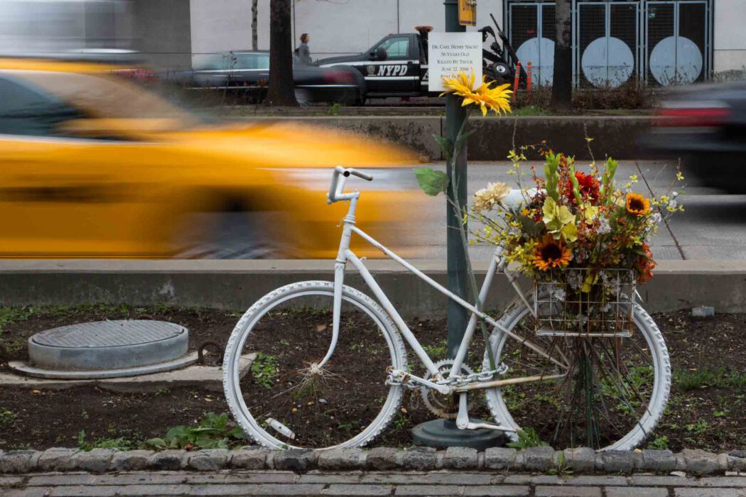 Weißes Ghost Bike als Gedenkstätte für einen getöteten Radfahrer in New York, mit Blumen geschmückt, im Hintergrund verschwommene gelbe Taxis und ein NYPD-Abschleppwagen.