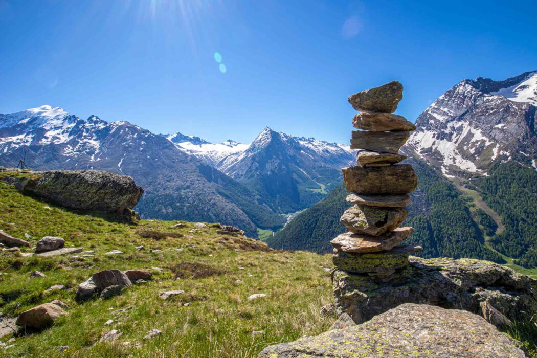 Steinmännchen / Steinturm auf Berggipfel mit Blick auf schneebedeckte Alpengipfel und grünes Tal bei strahlendem Sonnenschein.