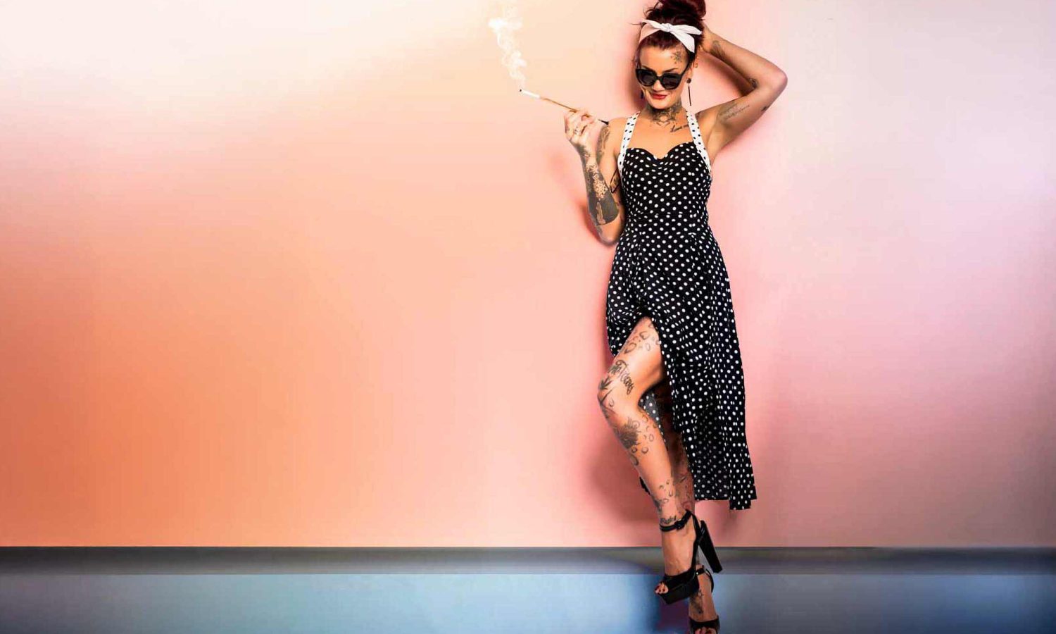 Ganzkörperaufnahme einer Frau im Vintage-Pin-up-Look: schwarzes Neckholder-Kleid mit weißen Punkten und Beinschlitz, Tattoos an Armen und Beinen, Sonnenbrille, Kopftuch und Zigarettenspitze, vor einer pastellrosa Wand mit dunklerem Bodenbereich