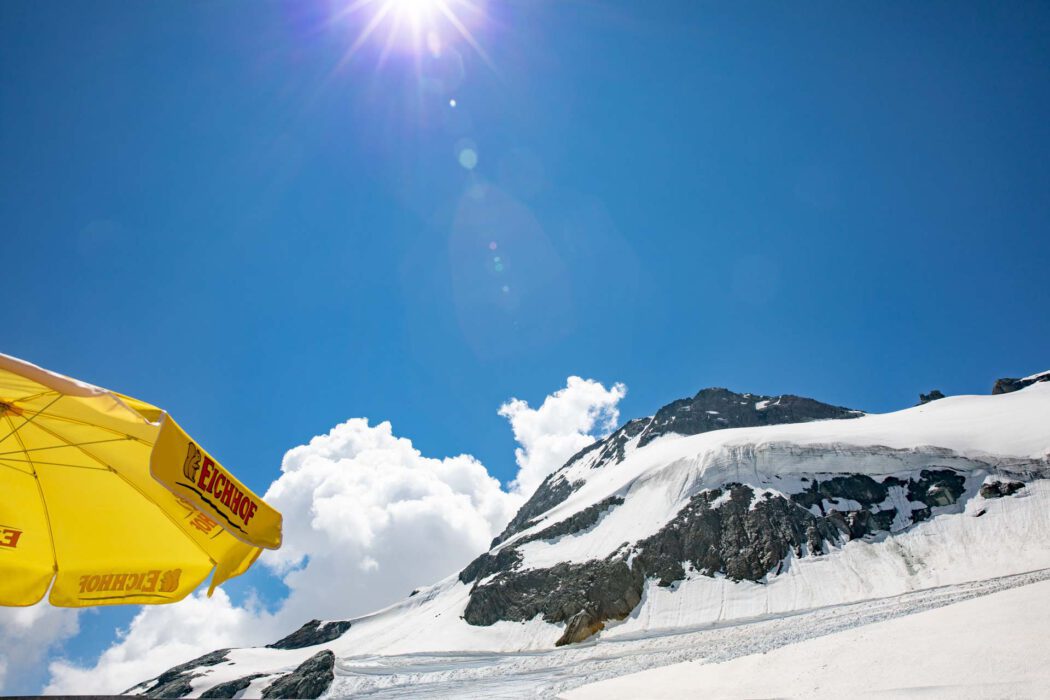 Reisefotografie Titlis Gletscher im Sommer: Schneebedeckter Hang mit Fels, strahlend blauer Himmel mit Sonne und Lens Flare, gelber Sonnenschirm mit Logo Eichhof am Rand, Skipiste, hochalpine Sommerstimmung.