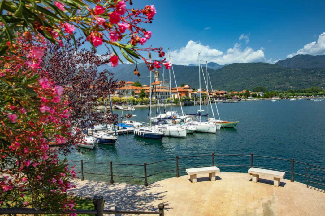 Kleiner Segelboothafen in Feriolo am Lago Maggiore mit weißen Yachten, im Vordergrund rosa Oleanderblüten und zwei Bänke auf einer Aussichtsterrasse, im Hintergrund Berge.