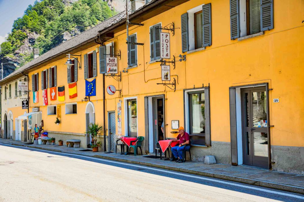 Gelbes Gebäude Ristorante Posta und Bar Posta an einer Dorfstraße im Piemont mit Flaggen von Italien, Schweiz, Deutschland und EU, Mann sitzt an einem Tisch mit roter Tischdecke vor dem Restaurant.