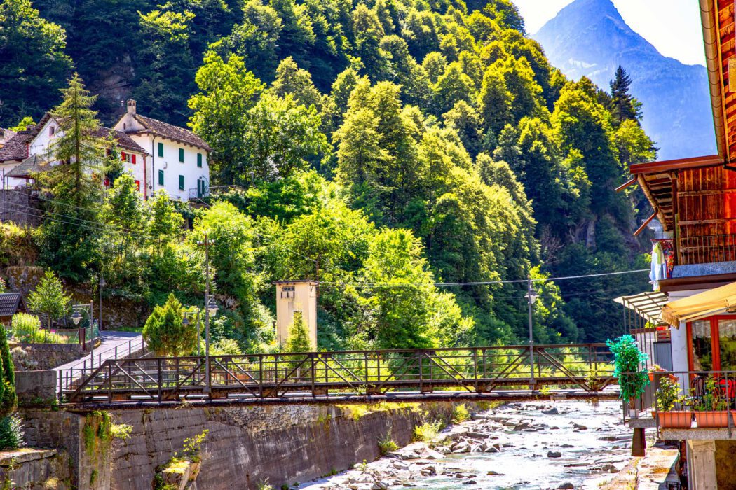 Kleine Fußgängerbrücke über einen Gebirgsfluss in einem italienischen Bergdorf im Piemont, umgeben von bewaldeten Bergen und historischen Häusern mit grünen Fensterläden