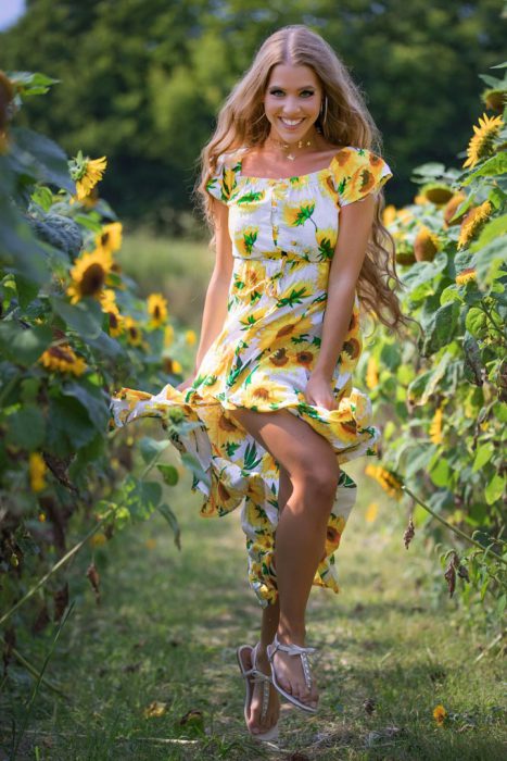 Eine lächelnde Frau mit langen, gewellten blonden Haaren trägt ein weißes Sommerkleid mit gelben Sonnenblumenmustern und steht in einem Feld voller hoher Sonnenblumen. Sie hat die Beine gekreuzt und ihre Füße schweben leicht über dem Boden, wobei weiße Riemchensandalen sichtbar sind.