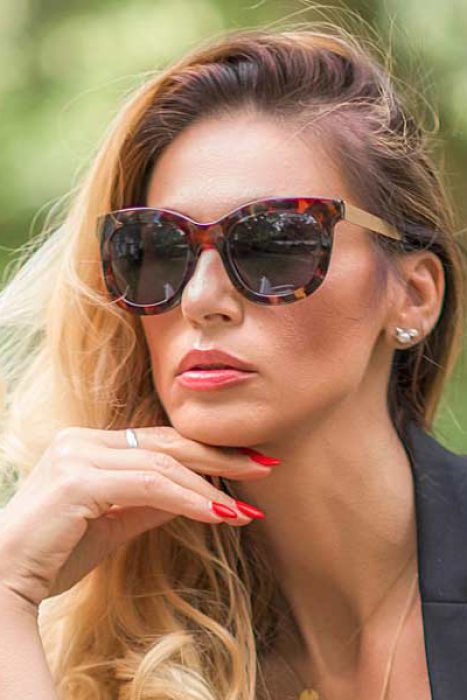 Nahaufnahme-Porträt einer Frau mit langer blonder, gewellter Haare und braun-gemusterter Sonnenbrille, die die Hand mit roten Nägeln am Kinn hält und roten Lippenstift trägt, mit unscharfem grünem Hintergrund.