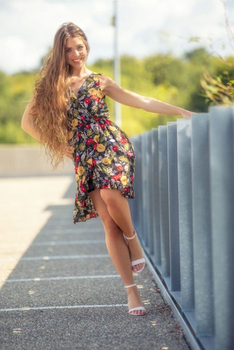 Eine lächelnde Frau mit langen, gewellten braunen Haaren trägt ein knielanges, schwarzes Sommerkleid mit buntem Blumenmuster (Rot, Gelb, Weiß). Sie lehnt sich entspannt an eine moderne, geriffelte graue Barriere im Freien und trägt weiße Riemchensandalen mit Absatz.