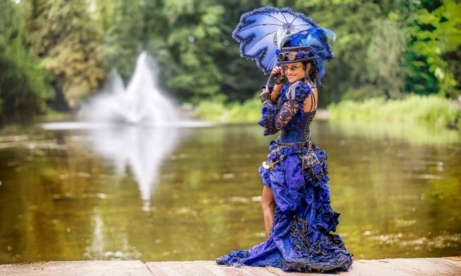 Am See – Zeitlose Eleganz – Steampunk Dame im royalblauen Rüschenkleid mit Federzylinder und Parasol posiert vor Fontäne am Parksee.