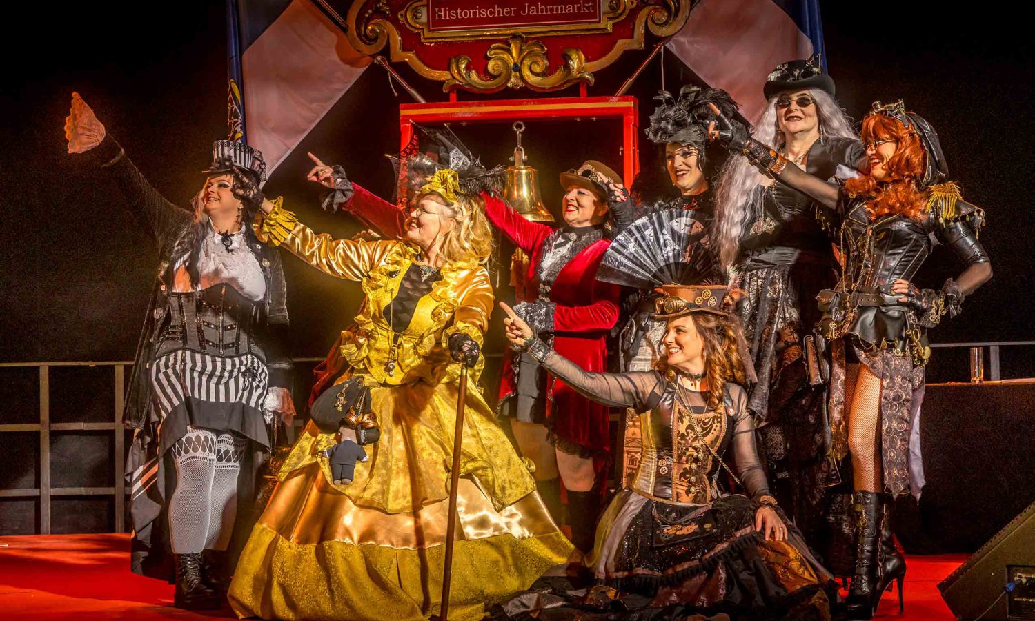 Acht Frauen in aufwendigen Steampunk Kostümen in Gold, Rot und Schwarz posieren lachend auf Bühne vor Schild Historischer Jahrmarkt mit Glocke, Abendveranstaltung mit Bühnenlicht – Steampunk Gruppe Eventfotografie.