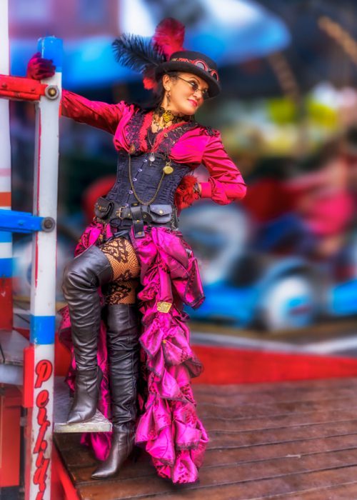 Eine Frau im Steampunk-Stil mit pinkem Rüschenrock, schwarzem Korsett, Stiefeln und einem Zylinder mit roten Federn steht auf einer Holzplattform.