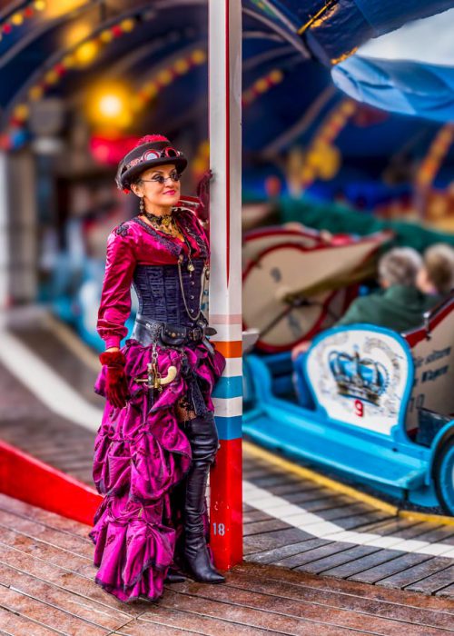 Eine Frau im kompletten Steampunk-Kostüm mit pinkem Rüschenrock, schwarzem Korsett, einem Zylinder mit Schutzbrille und Handschuhen steht an einem Pfosten. Im Hintergrund sind Fahrgeschäfte unscharf zu sehen.