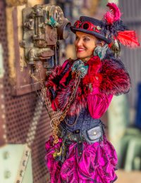 Eine lächelnde Frau in einem pinken und schwarzen Steampunk-Kostüm, komplett mit Zylinder mit roten Federn, flauschiger Stola, Korsett und Rüschenrock, hält einen Vintage-Telefonhörer an ihr Ohr und schaut zur Seite.