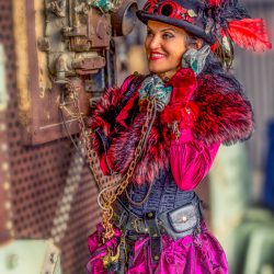 Eine lächelnde Frau in einem pinken und schwarzen Steampunk-Kostüm, komplett mit Zylinder mit roten Federn, flauschiger Stola, Korsett und Rüschenrock, hält einen Vintage-Telefonhörer an ihr Ohr und schaut zur Seite.