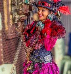 Eine lächelnde Frau in einem pinken und schwarzen Steampunk-Kostüm, komplett mit Zylinder mit roten Federn, flauschiger Stola, Korsett und Rüschenrock, hält einen Vintage-Telefonhörer an ihr Ohr und schaut zur Seite.