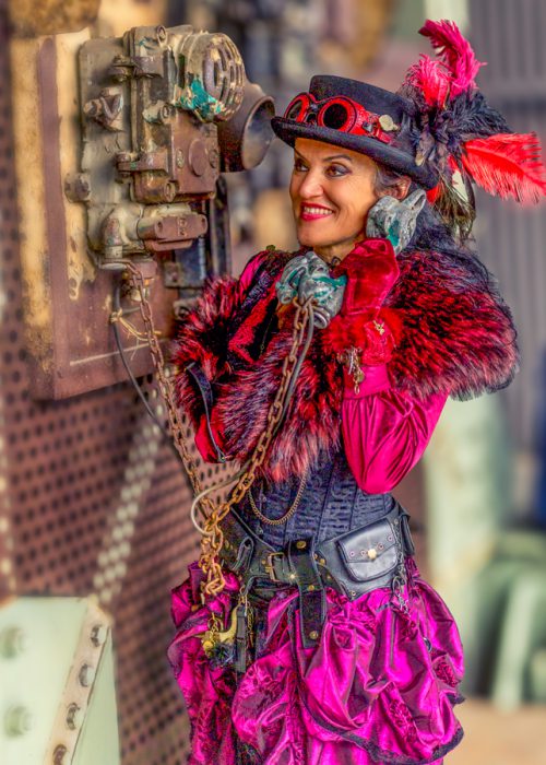 Eine lächelnde Frau in einem pinken und schwarzen Steampunk-Kostüm, komplett mit Zylinder mit roten Federn, flauschiger Stola, Korsett und Rüschenrock, hält einen Vintage-Telefonhörer an ihr Ohr und schaut zur Seite.