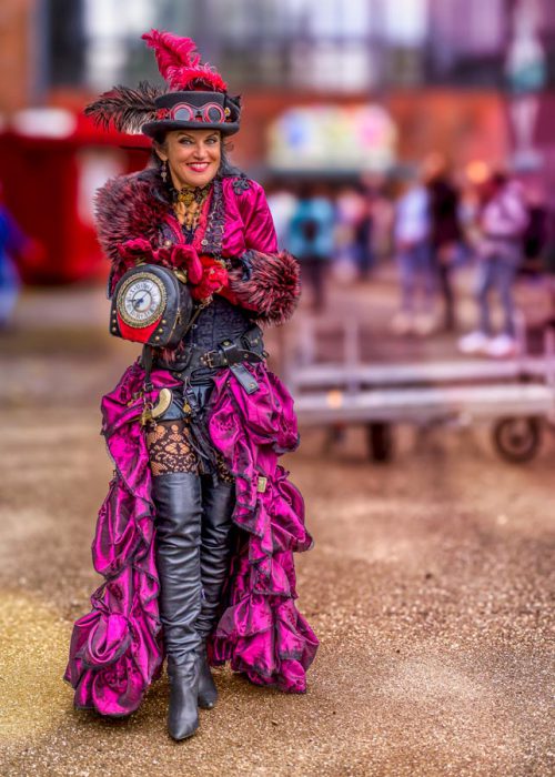 Eine lächelnde Frau in einem aufwendigen pinken und schwarzen Steampunk-Kostüm, bestehend aus einem Rüschenrock, Korsett, Stiefeln, einem Hut mit Federn und einer pelzigen Stola. Sie hält eine dekorierte Tasche mit einer großen Uhr in der Hand.