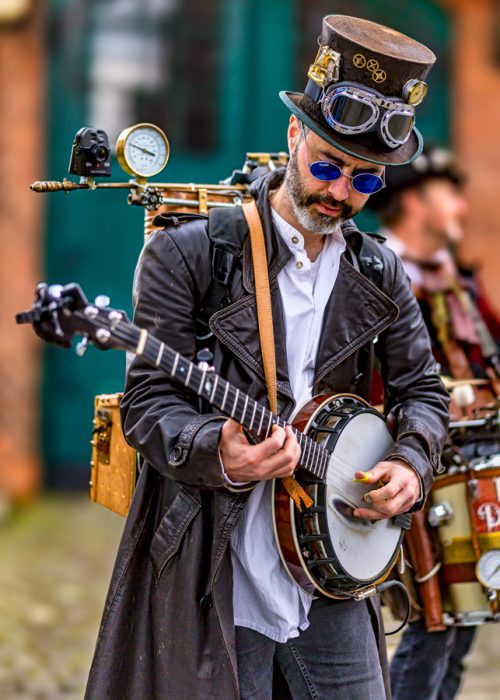 Ein Mann mit Bart, Sonnenbrille und Steampunk-Zylinder trägt eine braune Lederjacke und spielt ein Banjo. Auf seinem Rücken ist eine Apparatur mit Messgeräten und einer Kamera zu sehen.