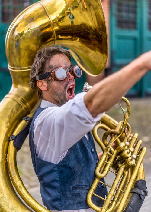 Ein Mann im Steampunk-Stil mit Schutzbrille und verschmiertem Gesicht spielt ein großes goldenes Sousaphon oder eine Tuba und hat den Mund weit geöffnet, als ob er singt oder ruft. Er trägt ein weißes Hemd und eine dunkelblaue Weste.