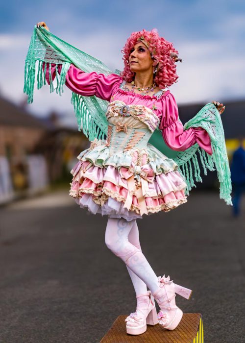 Eine Person mit pinker Lockenperücke und einem aufwendigen, mehrschichtigen Rüschenkleid in Pastellfarben (Pink, Hellblau, Creme) hält ein mintgrünes Fransen-Tuch über den Kopf. Sie trägt weiße Strumpfhosen mit Muster und pinke Plateau-Schuhe mit Blumen.