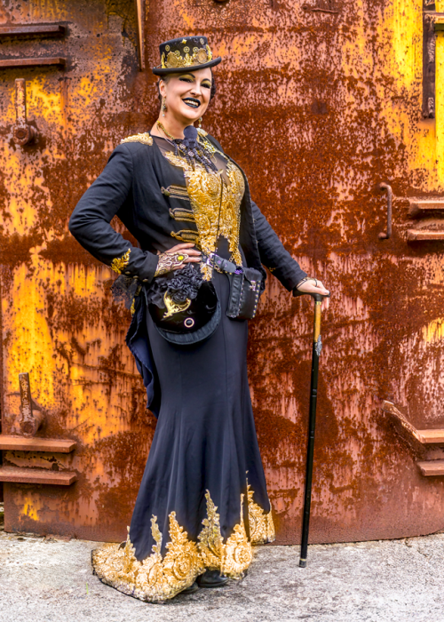 Eine Frau in einem eleganten schwarzen Steampunk-Kleid mit aufwendigen goldenen Stickereien und Verzierungen steht vor einer rostigen Metallwand. Sie trägt einen passenden Zylinder und lehnt sich auf einen Gehstock.