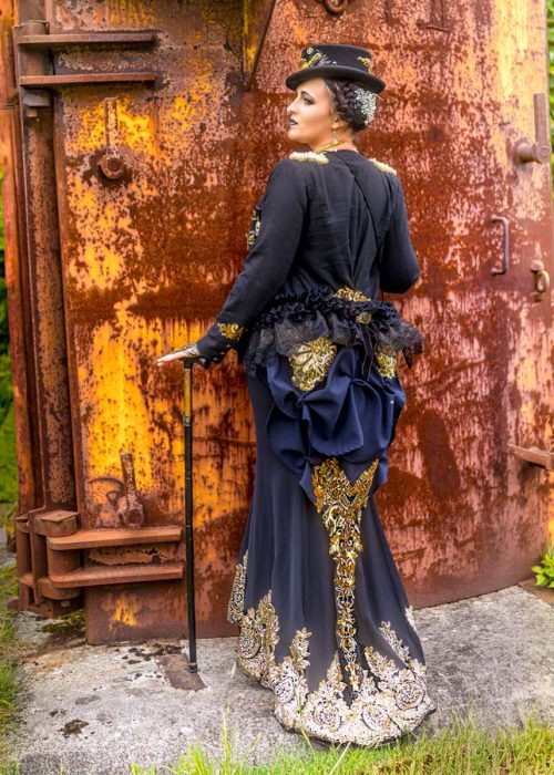 Rückenansicht einer Frau in einem eleganten schwarzen Steampunk-Kleid mit goldener Stickerei, einer blauen Raffung am Rücken und einem kleinen Zylinder. Sie steht vor einer rostigen Metallwand und hält einen Gehstock.