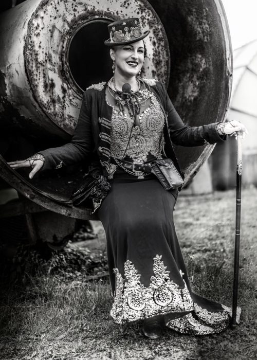 Eine Frau in einem kunstvollen Steampunk-Kleid mit reicher Stickerei und einem verzierten Zylinder sitzt lächelnd auf einer großen, verrosteten Metallröhre und hält einen Gehstock. Das Bild ist in Schwarz-Weiß gehalten.