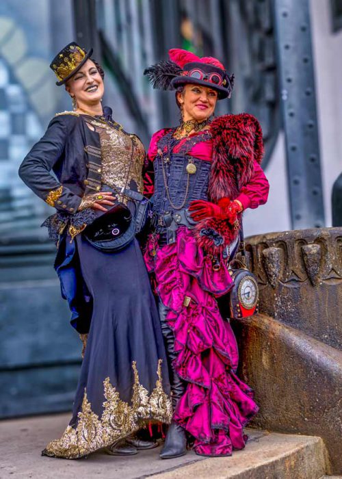 Zwei Frauen in detaillierten Steampunk-Kostümen stehen nebeneinander. Die linke Frau trägt ein schwarzes Kleid mit goldener Stickerei und einen Zylinder, während die rechte Frau ein pinkes Rüschenkleid, ein schwarzes Korsett, eine rote Stola und einen Hut mit Federn trägt. Im Hintergrund sind Industrieanlagen zu sehen.