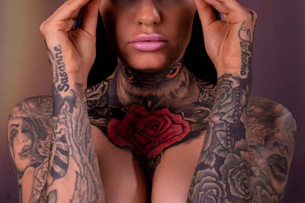 Nahaufnahme von Tattoo Model Denise, die eine pinke Baseballkappe trägt und diese mit beiden Händen festhält. Ihr Oberkörper und Hals sind mit großflächigen Tattoos bedeckt, darunter eine rote Rose auf der Brust und augenähnliche Motive am Hals. Auf ihren Händen sind ebenfalls Namen oder Wörter tätowiert. Sie blickt direkt in die Kamera.