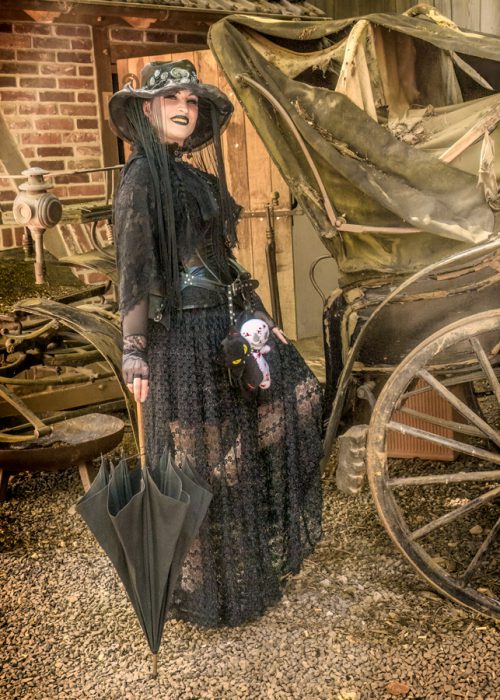 Eine Person in vollem Steam Punk Outfit, bestehend aus schwarzem Spitzenkleid, weitem Hut, Handschuhen und markantem Make-up, steht lächelnd neben einer historischen Kutsche. In der rechten Hand hält sie einen geschlossenen schwarzen Regenschirm und am Gürtel baumelt eine kleine schwarz-weiße Puppe. Im Hintergrund sind Teile eines Backsteinhauses zu sehen.