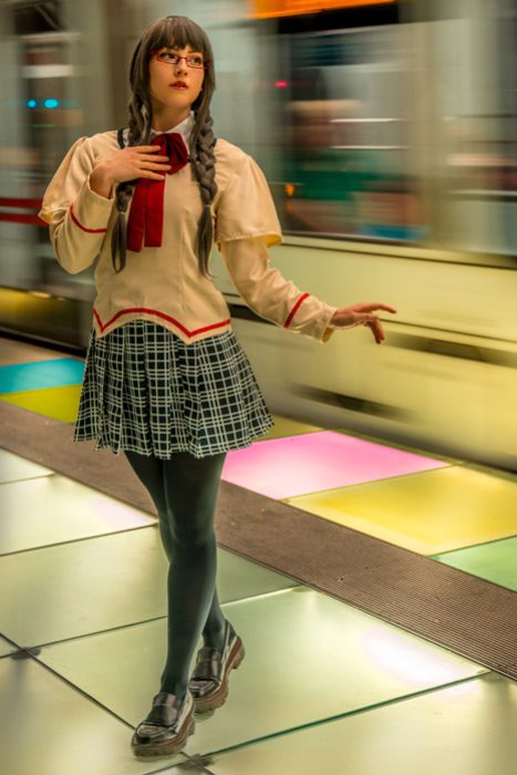 Eine Person in einer Schuluniform, bestehend aus einer beigen Bluse, einem karierten Rock und dunklen Strumpfhosen, steht auf einem U-Bahnsteig mit leuchtenden, farbigen Bodenplatten. Ein verschwommener Zug fährt im Hintergrund vorbei. Cosplay Fotoshootings
