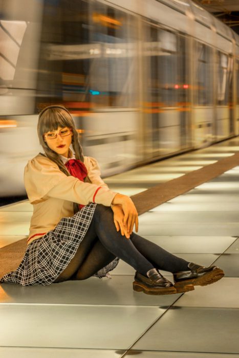 Eine Person in einer Schuluniform im Stil, bestehend aus einer beigen Bluse, einem karierten Rock und dunklen Strumpfhosen, sitzt auf einem U-Bahnsteig, während ein verschwommener Zug vorbeifährt. Cosplay Fotoshootings