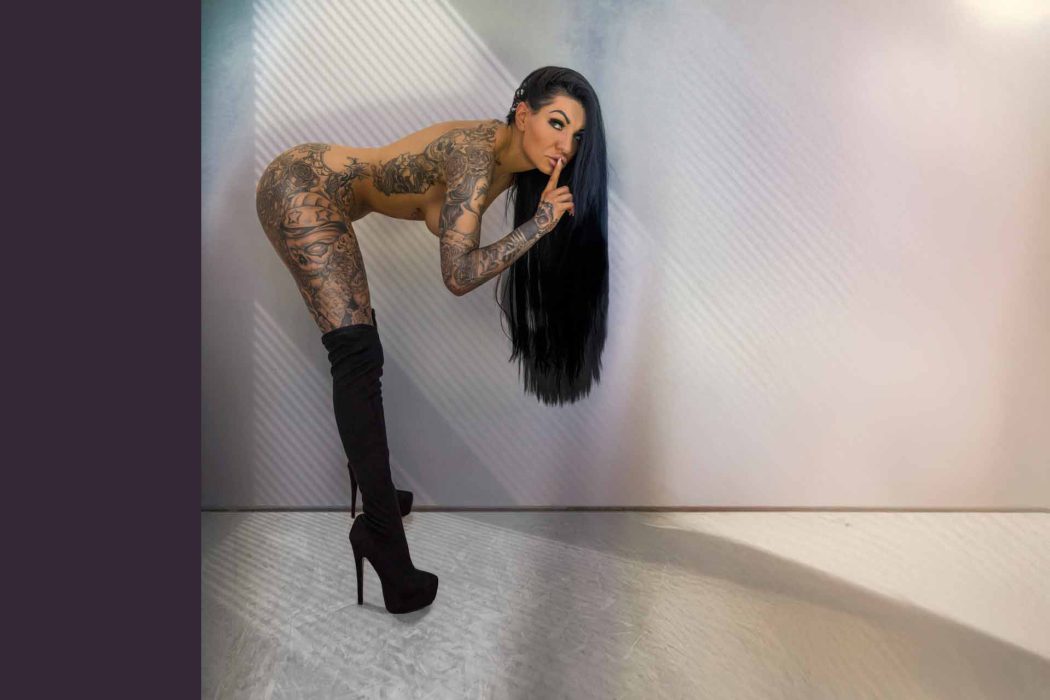 Eine Frau mit sehr langen, dunklen Haaren und großflächigen Tattoos auf den Armen lehnt sich von der Seite ins Bild. Sie hält einen Finger vor die Lippen, um zum Schweigen aufzufordern, und blickt direkt in die Kamera. Der Hintergrund ist hell und unscharf.