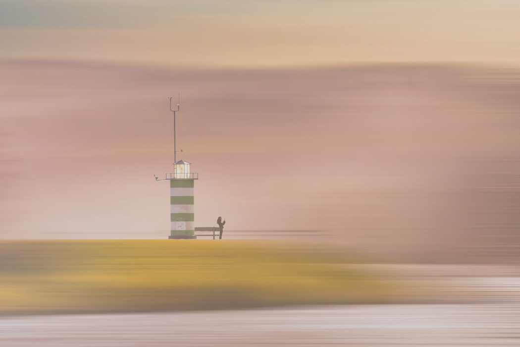 „Grün-weiß gestreifter Leuchtturm mit Person am Fuß vor pastellfarbenem Himmel bei Sonnenuntergang