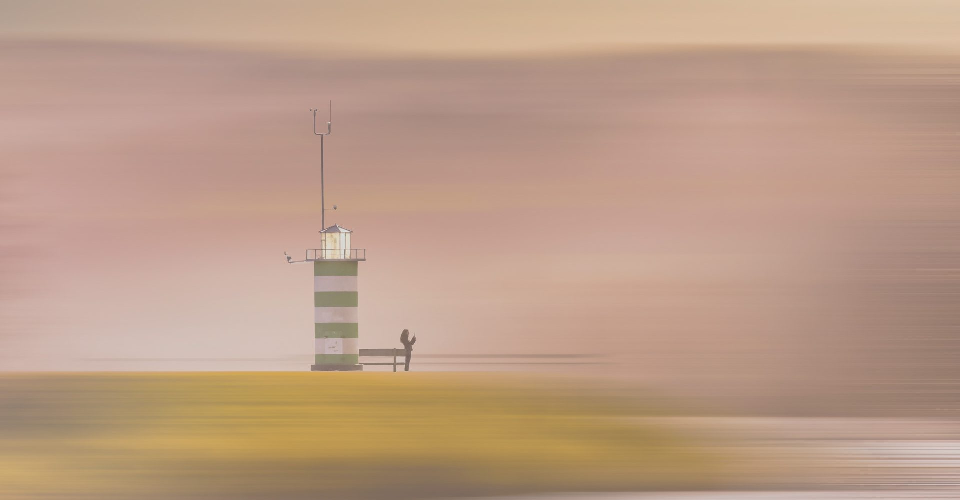 „Grün-weiß gestreifter Leuchtturm mit Person am Fuß vor pastellfarbenem Himmel bei Sonnenuntergang