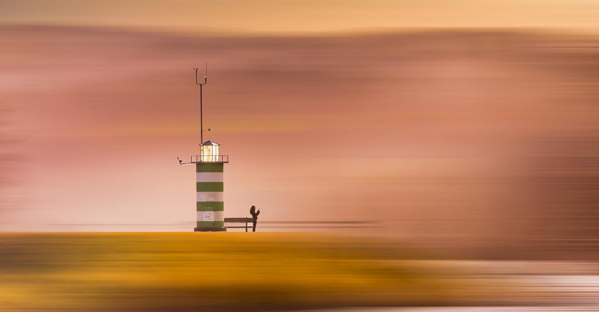 „Grün-weiß gestreifter Leuchtturm mit Person am Fuß vor pastellfarbenem Himmel bei Sonnenuntergang