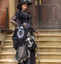 Frau in schwarzem Gothic Victorian Steampunk Kleid mit mehrlagigem Rüschenrock in Schwarz-Weiß, Spitze, Fransen, silbernen Ketten, großer Federhut und schwarz-weiß gestreiftem Gehstock, Tasche mit Totenkopf Plüschfigur, posiert auf verwitterter Steintreppe vor dunkler Holztür mit Backsteinwand – Steampunk Portrait Eventfotografie.