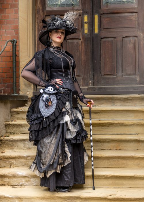 Frau in schwarzem Steampunk-Outfit mit Hut und Gehstock.
