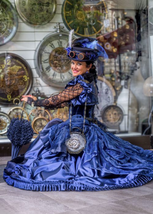 Frau in blauem Steampunk-Kleid vor Uhren-Schaufenster.