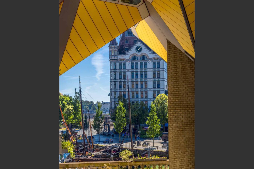Reisefotografie Rotterdam: Blick durch die gelben Kubuswoningen Kubushäuser auf das historische Witte Huis Weißes Haus mit Uhrturm am Oude Haven Alter Hafen, Segelschiffe, Bäume und Cafés im Sonnenschein, architektonischer Kontrast.