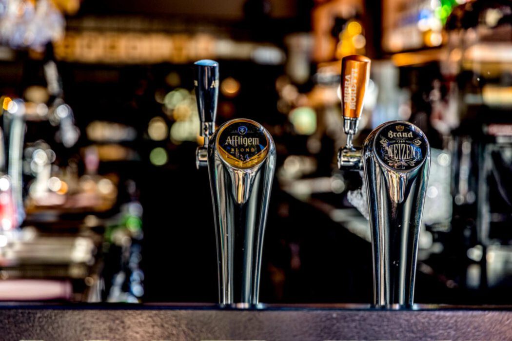 Reisefotografie Bar in Rotterdam: Zwei glänzende Bier-Zapfhähne Nahaufnahme – Affligem Blond und Brand Weizen 1340, im Hintergrund Birra Moretti, unscharfer Kneipen-Hintergrund mit warmem Bokeh, Theken-Detail.