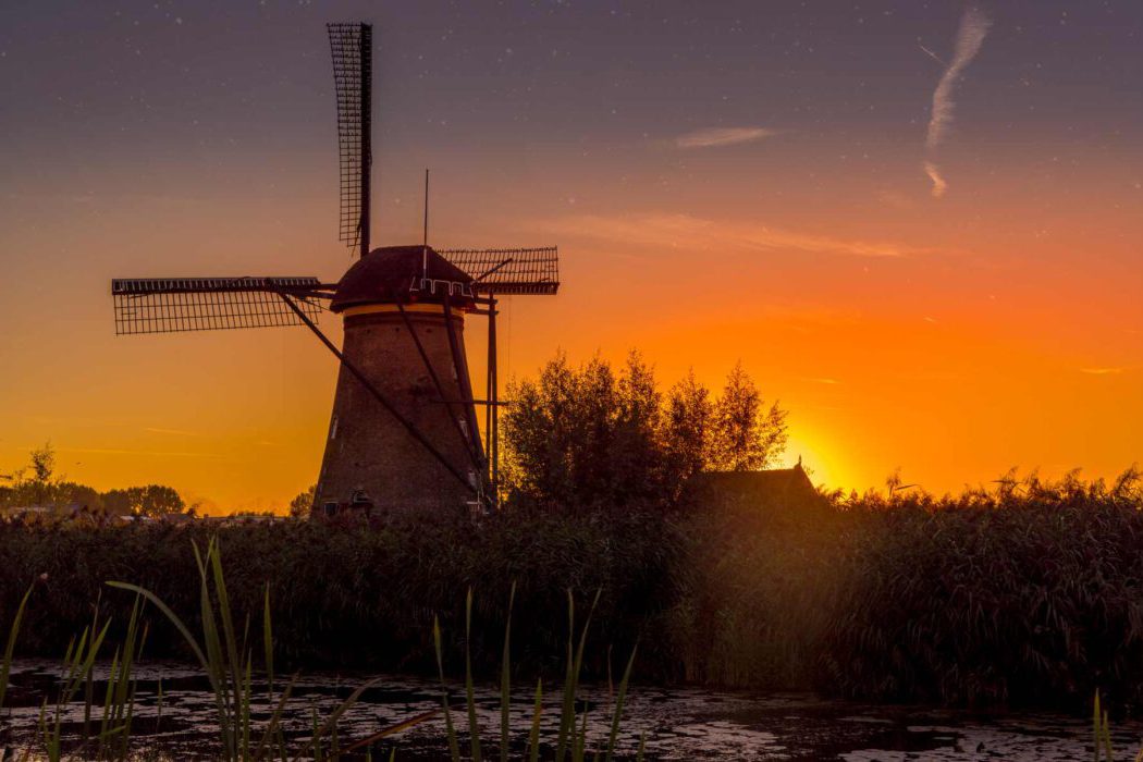 Windmühle Kinderdijk Reisefotografie Kinderdijk bei Sonnenuntergang: Historische Backstein-Windmühle als Silhouette vor intensiv orange-gelbem Himmel, Sonne geht hinter Bäumen unter, Schilf und Kanal im Vordergrund, dramatisches Abendlicht.