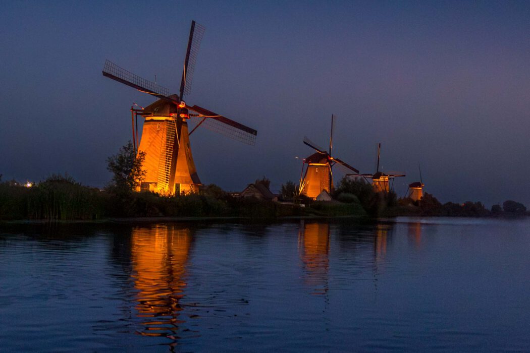 Vier illuminierte Windmühlen von Kinderdijk bei Nacht am Kanal, warm gold beleuchtet, Spiegelung im ruhigen Wasser, blaue Stunde, UNESCO Welterbe Kinderdijk Niederlande, Nacht- und Reisefotografie.